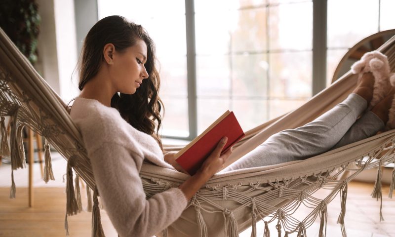 Dark-haired girl dressed in pants, sweater and warm slippers reads a book lying in a hammock in a cozy room with panoramic windows
